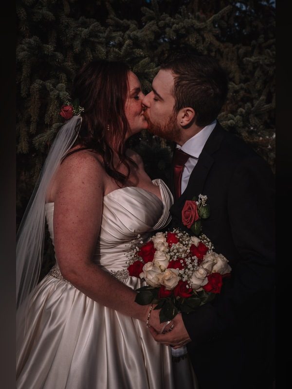Bride and groom kissing in front of an evergreen tree, holding a bouquet of red and white roses