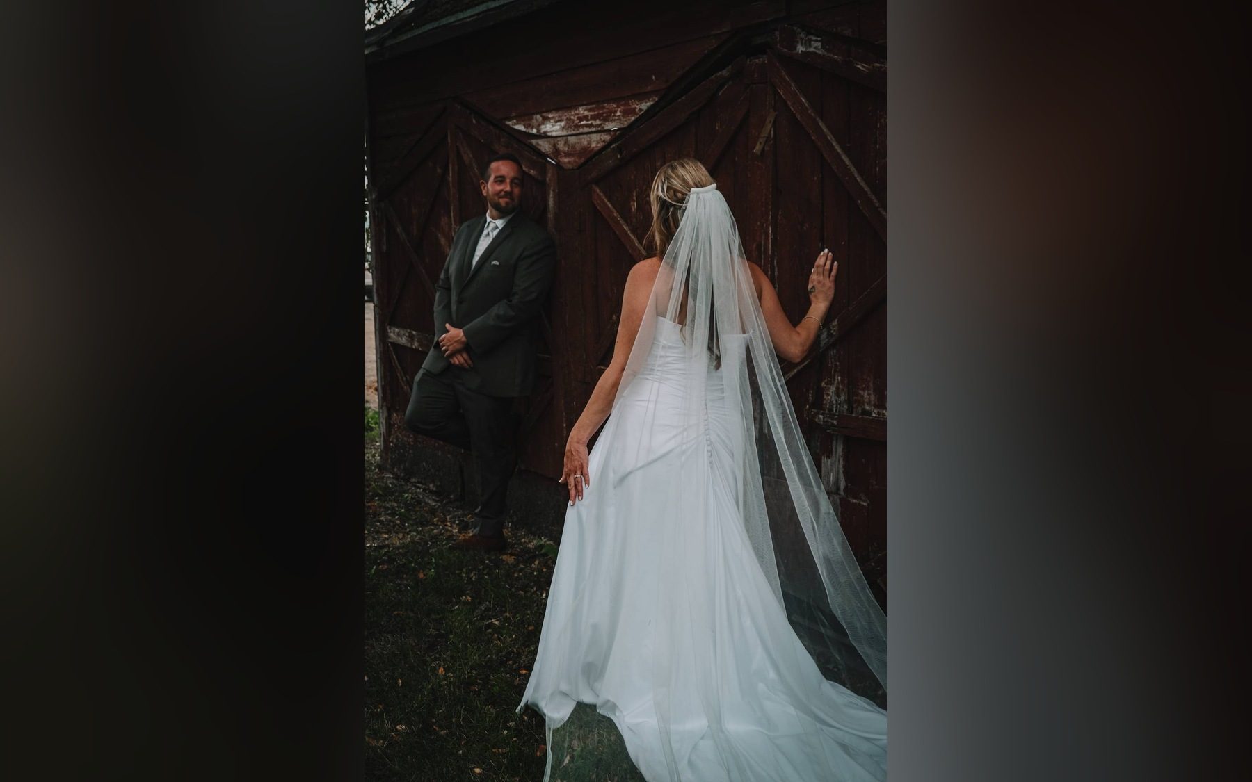 Groom leaning against an old weathered barn door as the bride in a long veil walks toward him