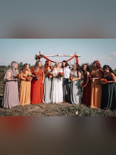 Bride and groom with bridesmaids in autumn-colored dresses holding sunflower bouquets under a wooden arch