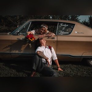 Couple posing by a vintage car, one leaning out of the window and the other sitting in the grass