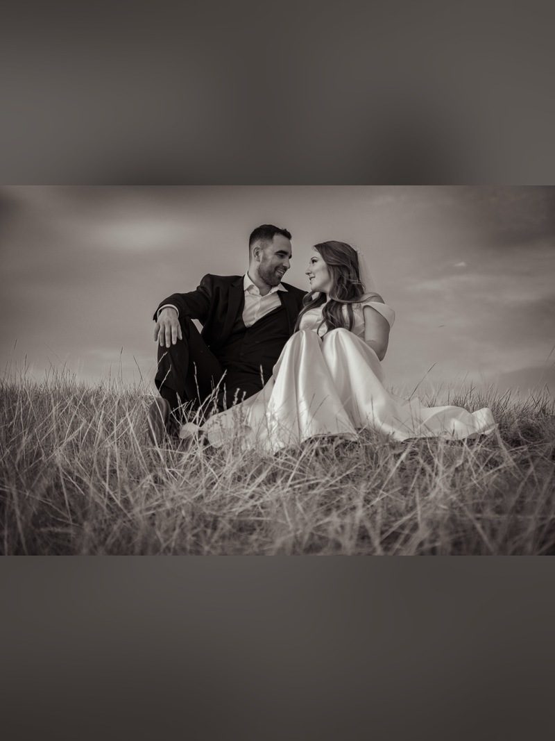 Bride and groom sitting in tall grass looking at each other under a cloudy sky, black and white