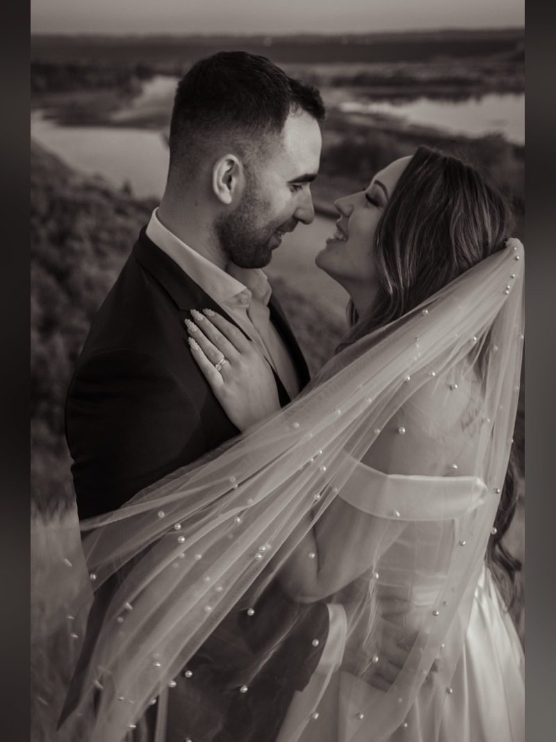 Couple embracing by a river, the bride wearing a pearl-studded veil, black and white