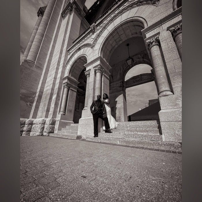 Bride and groom standing on the steps of a stone building with tall arches, black and white